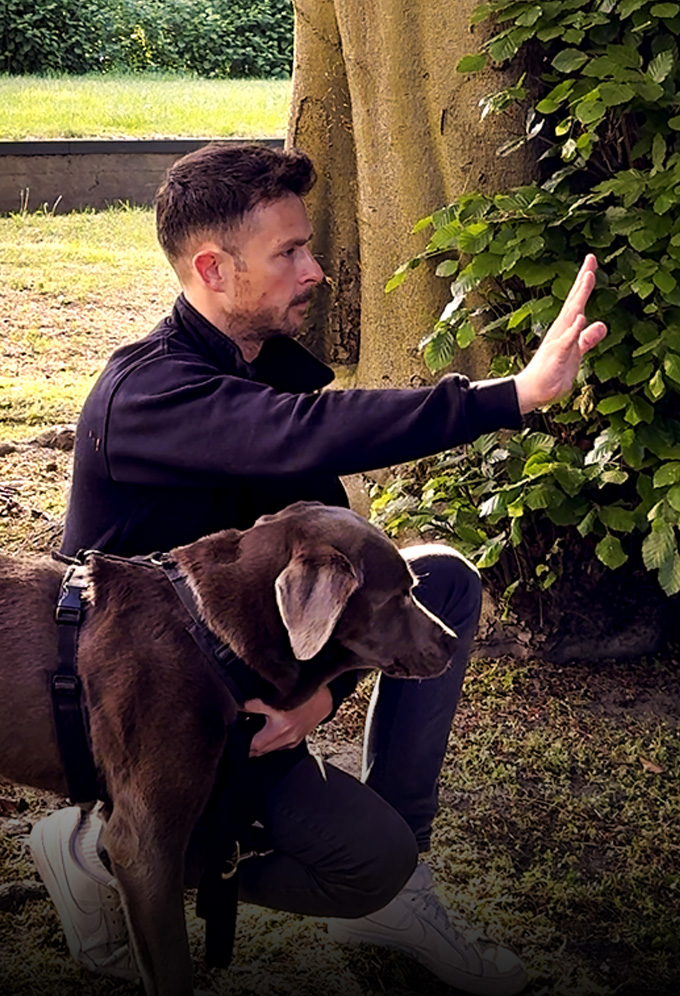 Ein Mann hockt neben seinem Hund und hebt ruhig eine Hand in Richtung anderer Menschen, um ein Signal zu geben. Der Hund steht aufmerksam an seiner Seite und schaut in die gleiche Richtung. - Rubrik Körpersprache Mensch und Hund - RUDEL. App
