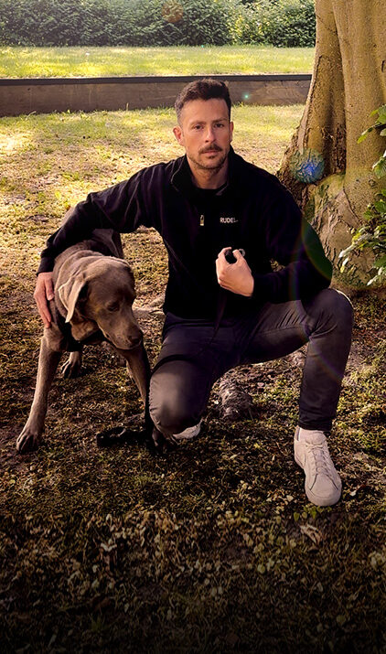 Ein Mann hockt neben seinem Hund und hebt ruhig eine Hand in Richtung anderer Menschen, um ein Signal zu geben. Der Hund steht aufmerksam an seiner Seite und schaut in die gleiche Richtung. - Rubrik Körpersprache Mensch und Hund - RUDEL. App