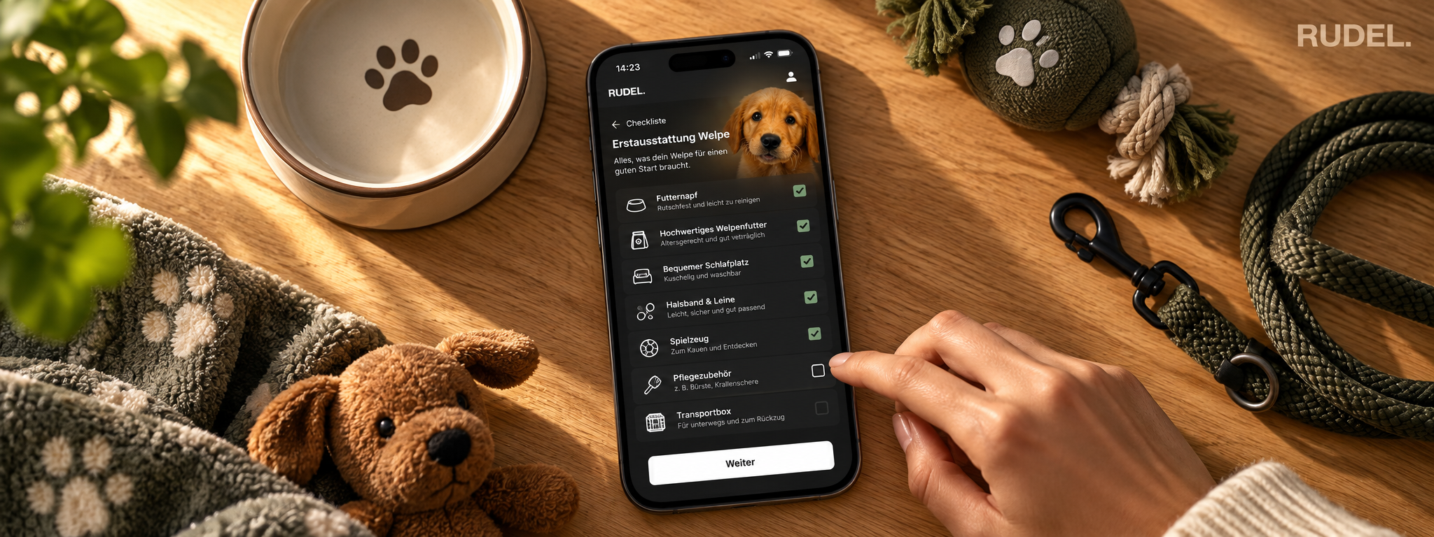 Eine Hand tippt auf einem Smartphone die Erstausstattungs-Checkliste in der RUDEL App durch, während Napf, Leine, Spielzeug und eine weiche Decke griffbereit auf dem Tisch liegen