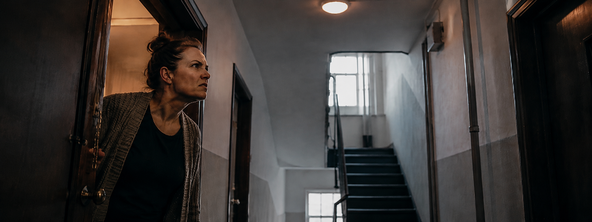 Frau schaut wachsam aus ihrer Wohnungstür in den Hausflur, im Hintergrund ein Treppenhaus